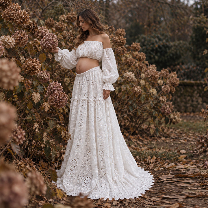 A woman wearing a white off-the-shoulder boho eyelet maxi dress stands outdoors in a garden path surrounded by autumn foliage and dried flowers.