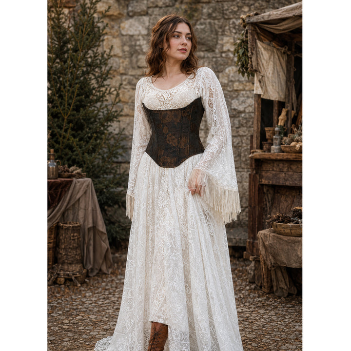 A woman stands before a stone wall wearing a Beroe beige lace gown with a brown corset. Her outfit has fringed wide cuffs and a brown boot.