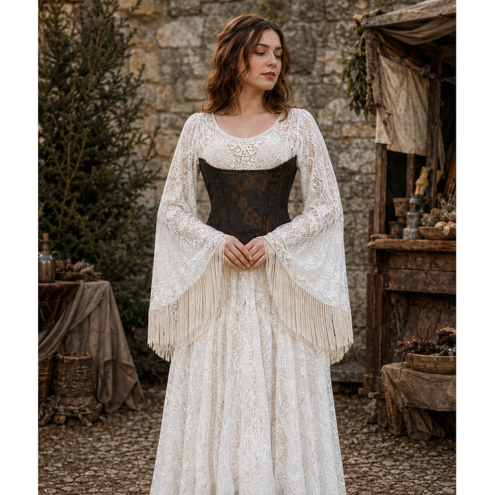 A woman stands before a stone wall wearing a Beroe beige lace gown with a brown corset. Her outfit has fringed wide cuffs and a brown boot.