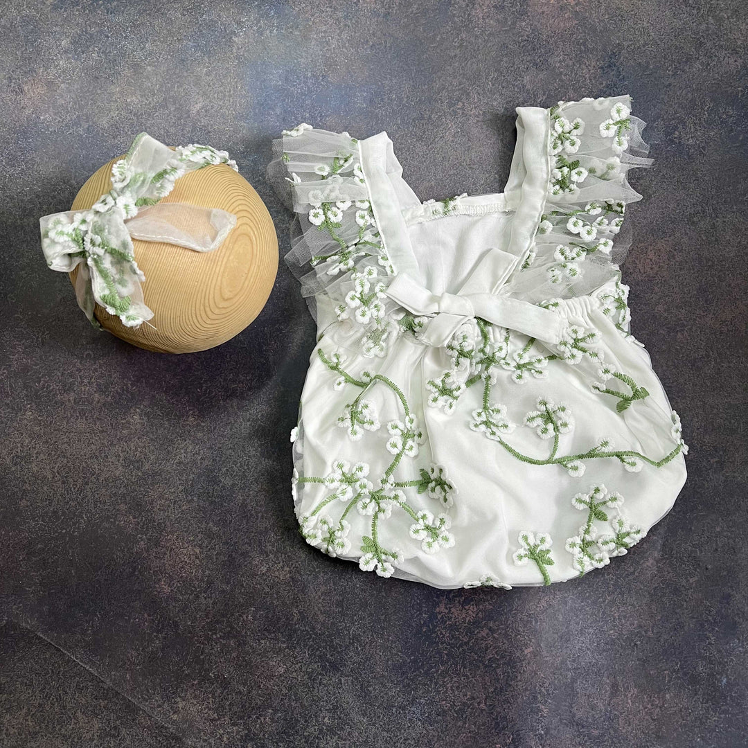 A romper laid flat from the back on a dark surface, with a bow tie at the waist and the same tulle flower headband on the left.