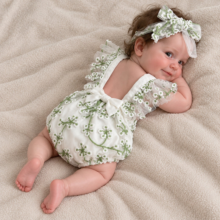 A baby in a white suspender romper with green floral embroidery and a white bow on the back, lying on her side on a beige blanket, wearing a matching tulle flower headband, head turned to the camera.