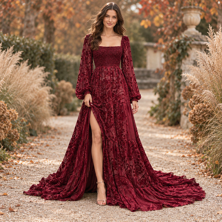 A woman in a Calypso red wine long-sleeve slit dress with heeled sandals stands on a gravel path with fallen leaves, dried grass and trees on both sides.