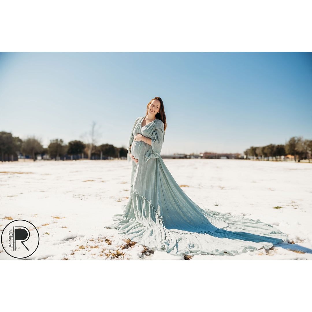 A pregnant woman in a Daphne light blue slit dress with a shawl stands on snow-covered grass, holding her belly.
