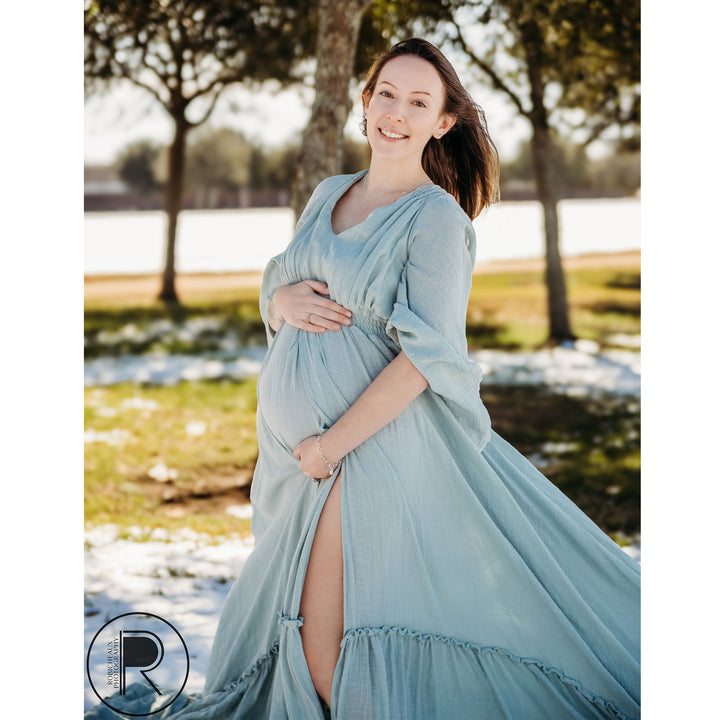 A pregnant woman in a light blue long-sleeve slit dress stands on snowy grass, hands on her belly.