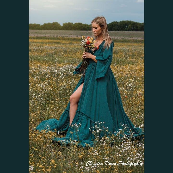 A woman in a Daphne emerald dress stands in a wildflower field, holding flowers in one hand and touching her hair with the other. The dress has a V-neck, flared sleeves, and a high slit.