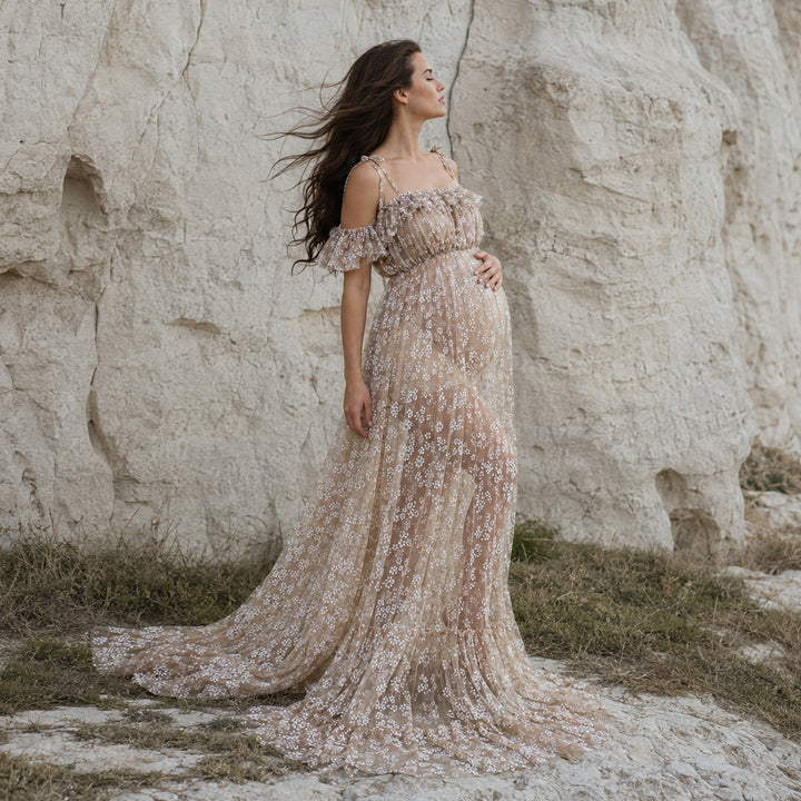 A pregnant woman stands before a Ersa khaki rock face wearing a sheer, floral embroidered off-shoulder gown.