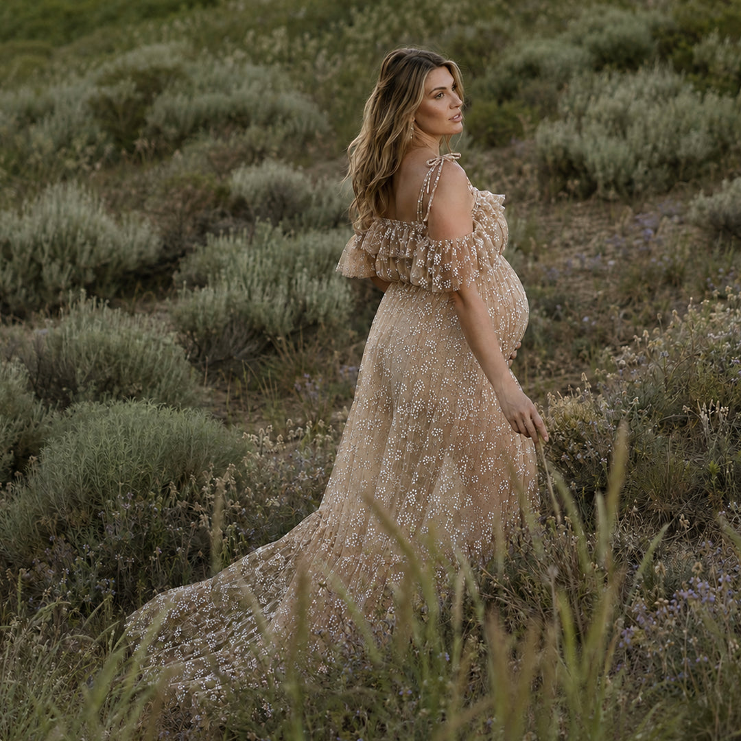 A pregnant woman stands in shrubs, wearing a Ersa khaki sheer, off-shoulder gown with ruffles.