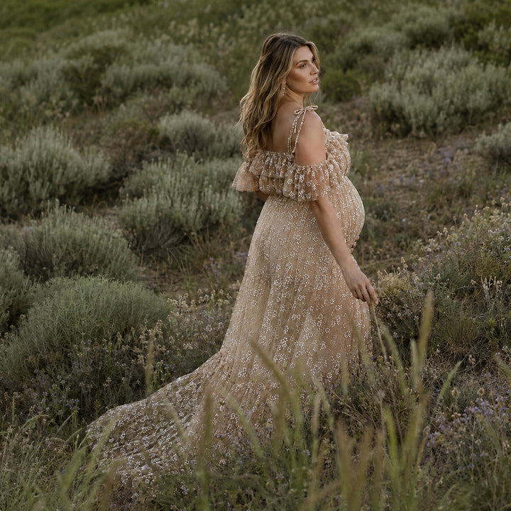 A pregnant woman stands in shrubs, wearing a Ersa khaki sheer, off-shoulder gown with ruffles.