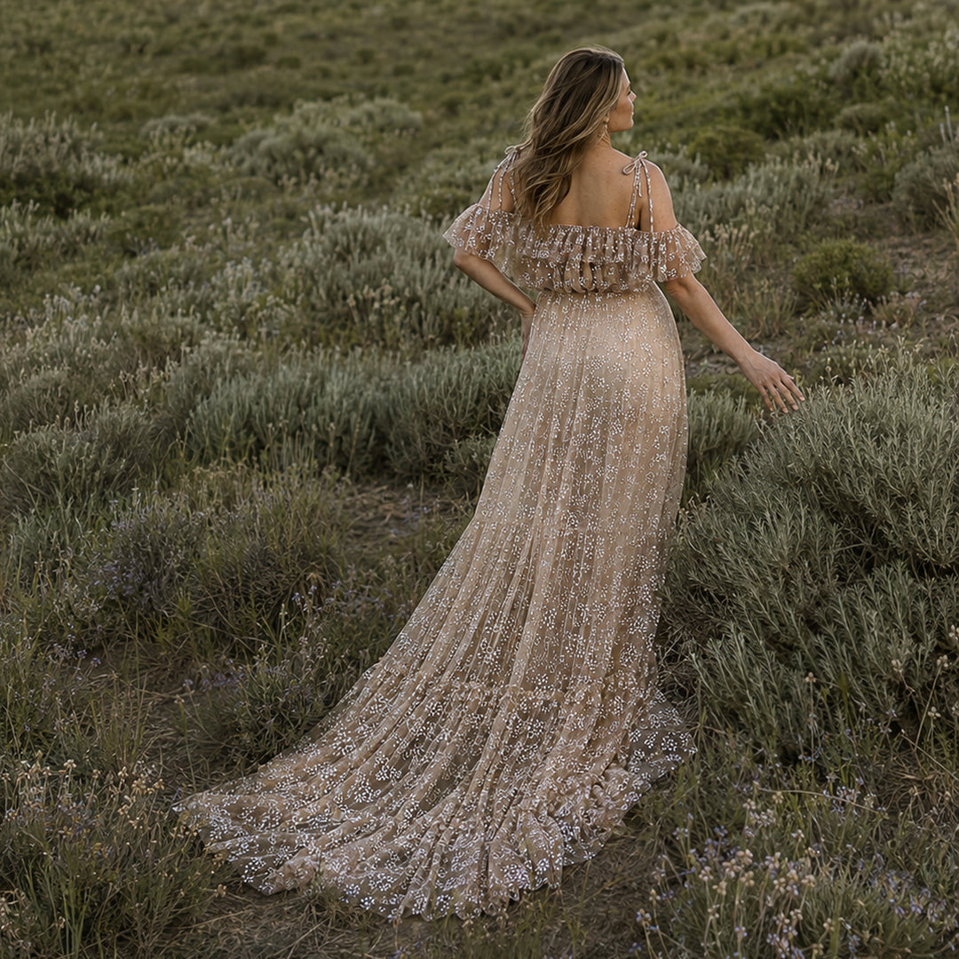 A pregnant woman stands in shrubs, wearing a Ersa khaki sheer, off-shoulder gown with ruffles.