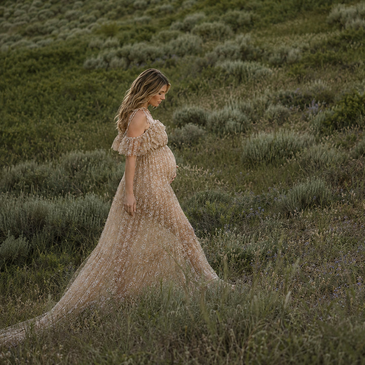 A pregnant woman stands in shrubs, wearing a Ersa khaki sheer, off-shoulder gown with ruffles.