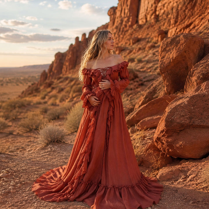 A woman stands outdoors in a desert canyon wearing a long, off the shoulder kallectoria terracotta 
gown with ruffled details and a full skirt 