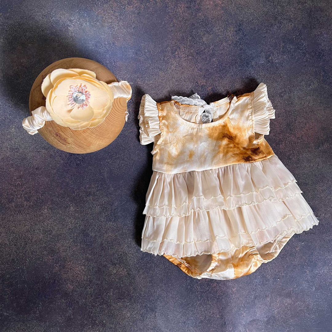 A romper laid flat from the front, showing the yellow tie-dye bodice, ruffled sleeves and three-tier skirt, with the same flower headband on the left.