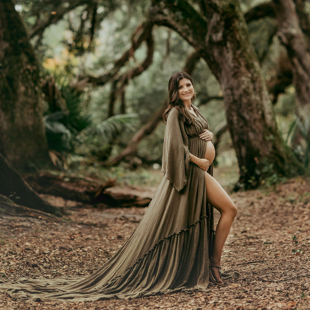 a pregnant woman in a Daphne olive long dress with a slit and shawl stands on leaf-covered forest ground, holding her belly.