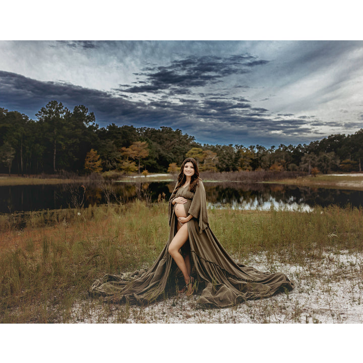 A pregnant woman in a Daphne olive long dress with a slit and shawl stands on grass by a pond, holding her belly.